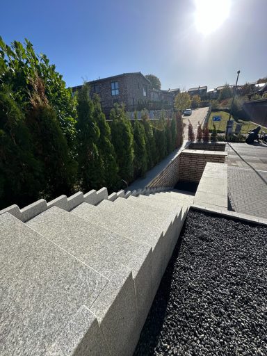 Steinige Treppe zu einem Garten mit Hecken und einem sonnigen Himmel im Hintergrund.