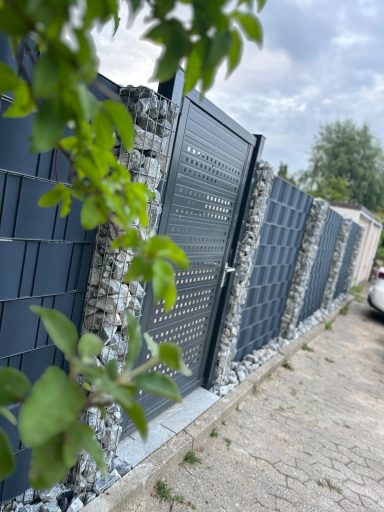 Grauer Zaun aus Stein mit metallener Tür, umgeben von grünem Laub.